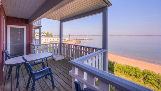 A covered deck with a table and chairs, overlooking a calm body of water and a distant shoreline.