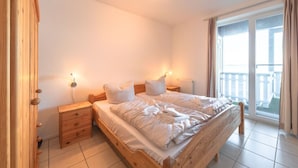 A bedroom with two beds, wooden headboards, bedside tables, and a view of a balcony through a large window.