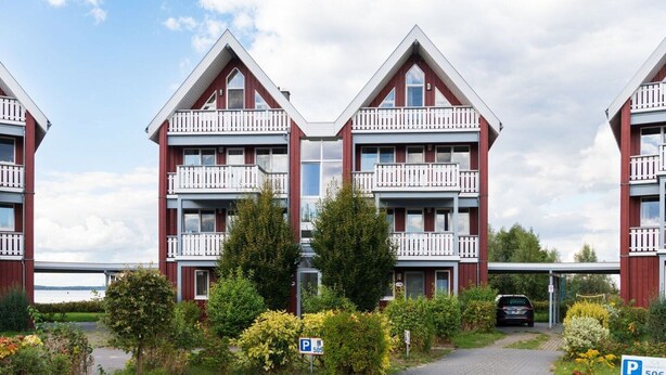 A row of red and white two-story apartment buildings with balconies, a paved driveway, and a parking area marked with a 'P' sign. - liebevoll eingerichtetes Appartement (Rechlin)