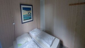 Ett litet rum med en enkel säng, en väggpanel av trä och en inramad tavla som föreställer ett landskap.