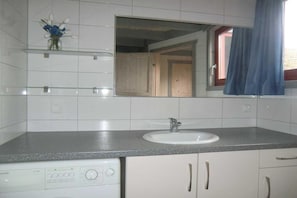 Un baño con un lavamanos blanco, un espejo encima y una repisa de cristal con un jarrón de flores.