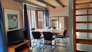 Ett rum med ett träpanelat tak, ett stort fönster med gardiner, en platt-TV på ett träställ, ett matbord med stolar och en bokhylla.