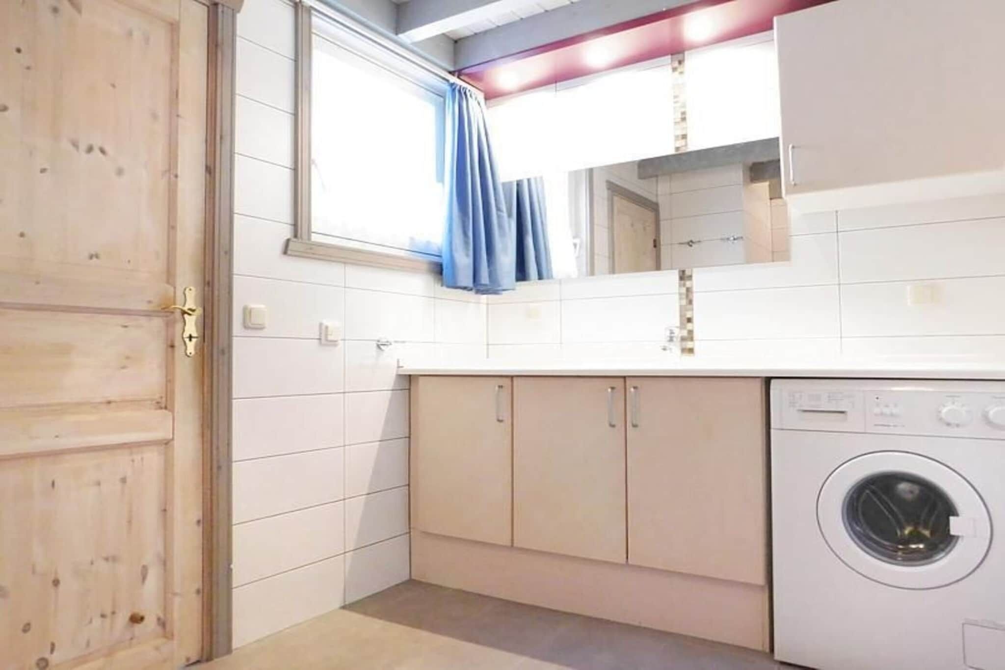 A compact kitchen with a built-in washing machine, wooden door, and a window with a blue curtain.