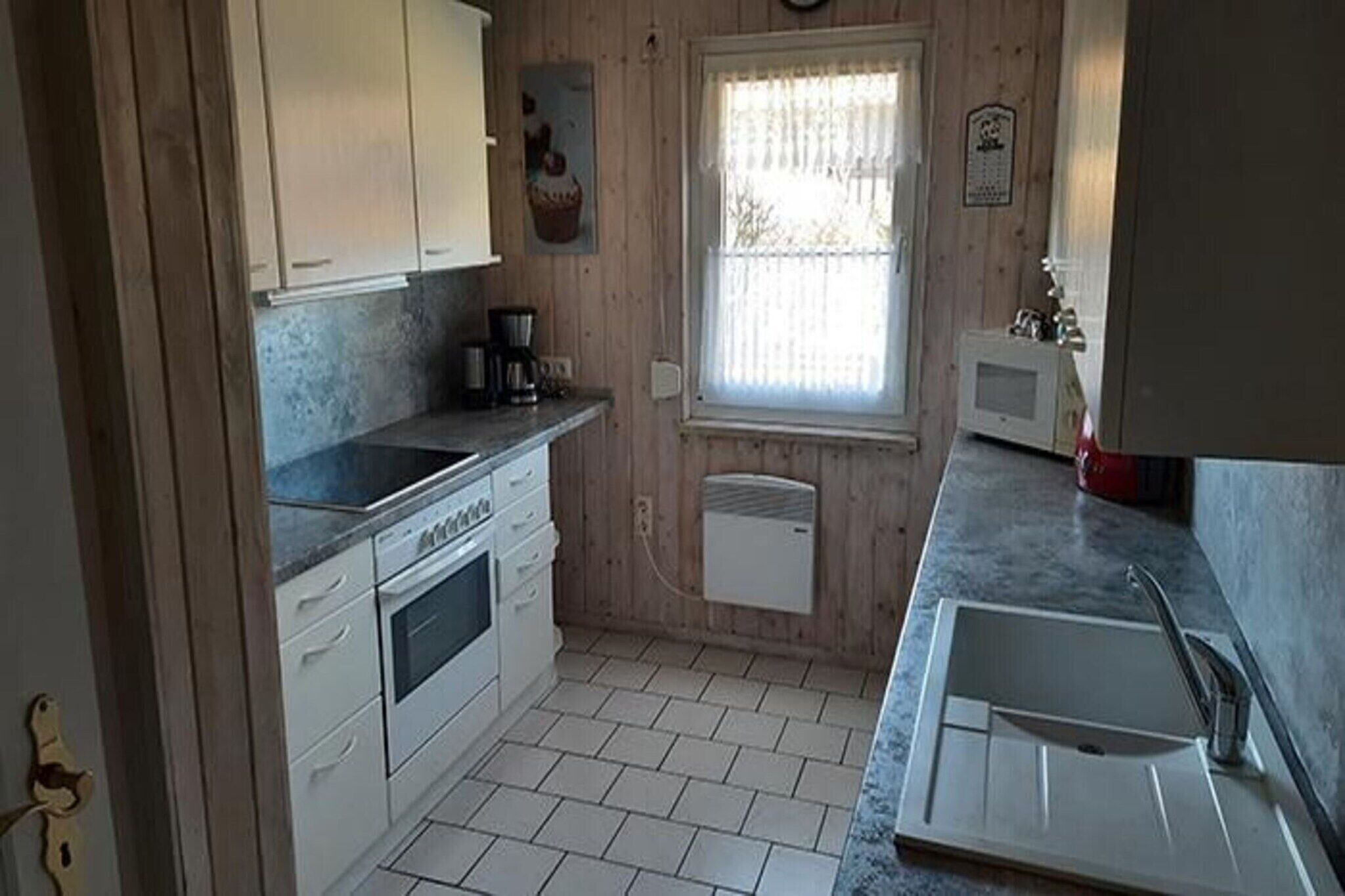 Uma cozinha compacta com armários brancos, forno embutido, cafeteira e janela com cortinas.