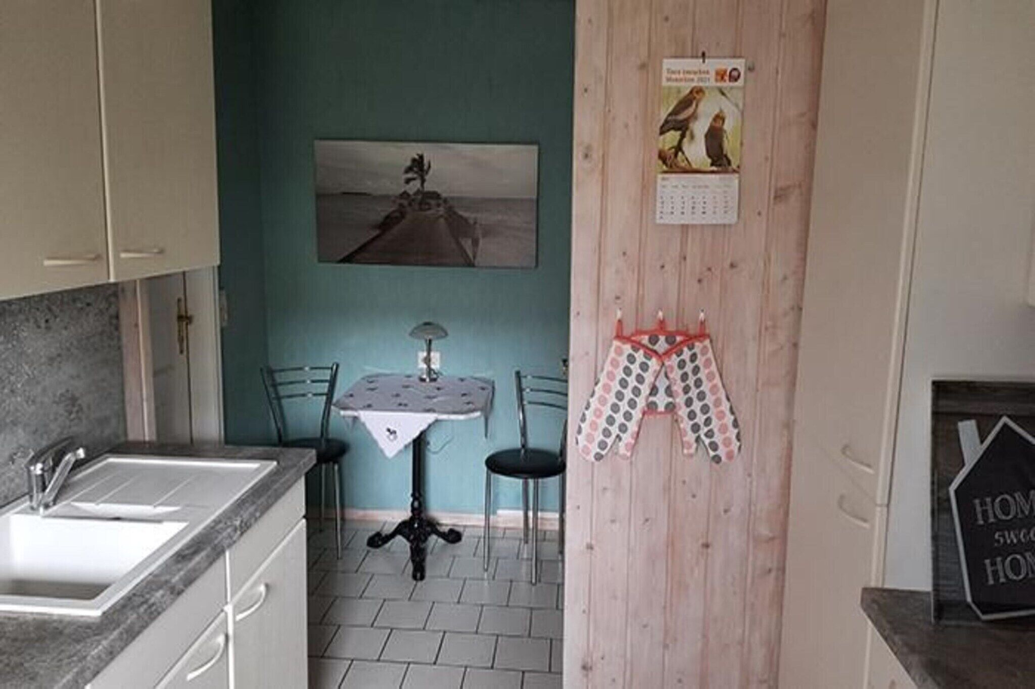 Et køkken med vask, et lille bord og stole. Der er en kalender og et skilt på væggen.