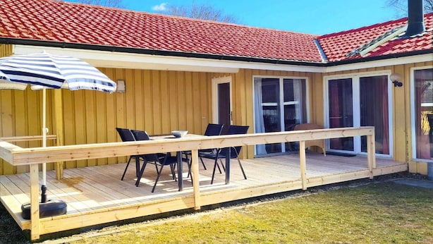 Een houten terras met een tafel, stoelen, een gestreepte parasol en een dak met rode tegels.