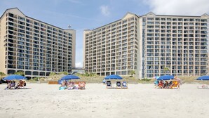 A beachfront hotel with multiple high-rise buildings, beach umbrellas, and lounge chairs.