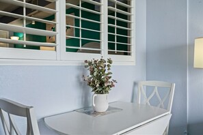 A white table with a vase of flowers, a white chair, and a window with white blinds. - Oceanfront Sunrise Condo with Heated Pools Hot Tub (North Myrtle Beach)