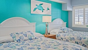 A room with two beds, a nightstand with a lamp, a wall painting of a turtle, and a window with blinds.