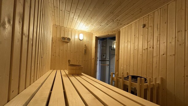 Una sauna de madera con bancos y un asiento integrado, un termómetro fijado a la pared y una puerta que da a otra habitación.