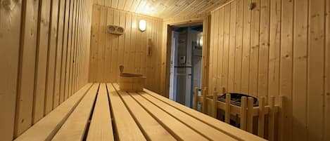 A wooden sauna with benches and a bench seat, a wall-mounted thermometer, and a door leading to another room.
