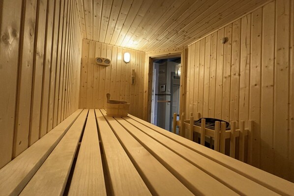 A wooden sauna with benches and a bench seat, a wall-mounted thermometer, and a door leading to another room.