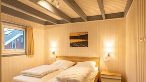 Cama doble con sábanas blancas, cabecera de madera, dos mesitas de noche, lámpara de pared y una ventana con cortinas.