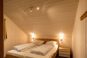 A room with wooden panel walls, a sloped ceiling, two beds with white bedding, and two wall-mounted light fixtures.
