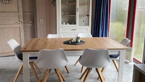 A dining area with a wooden table and white chairs, a cabinet with glass doors, and a view through a window with curtains. - skandinavisches Ferienhaus in Mirow (Granzow)
