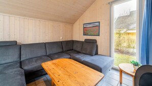 Ein modernes Wohnzimmer mit einem Ecksofa, einem Holztisch und einem großen Fenster mit Blick ins Freie.