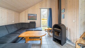 Ein gemütliches Zimmer mit Holzbalkendecke, einem grauen Sofa, einem Holztisch, einem kleinen runden Tisch mit Pflanze und einem Kaminofen.