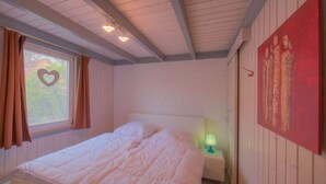 A bedroom with a bed, a nightstand, a window with curtains, and a wall with a painting. - Ferienhaus im Ferienpark Mirow (Granzow)