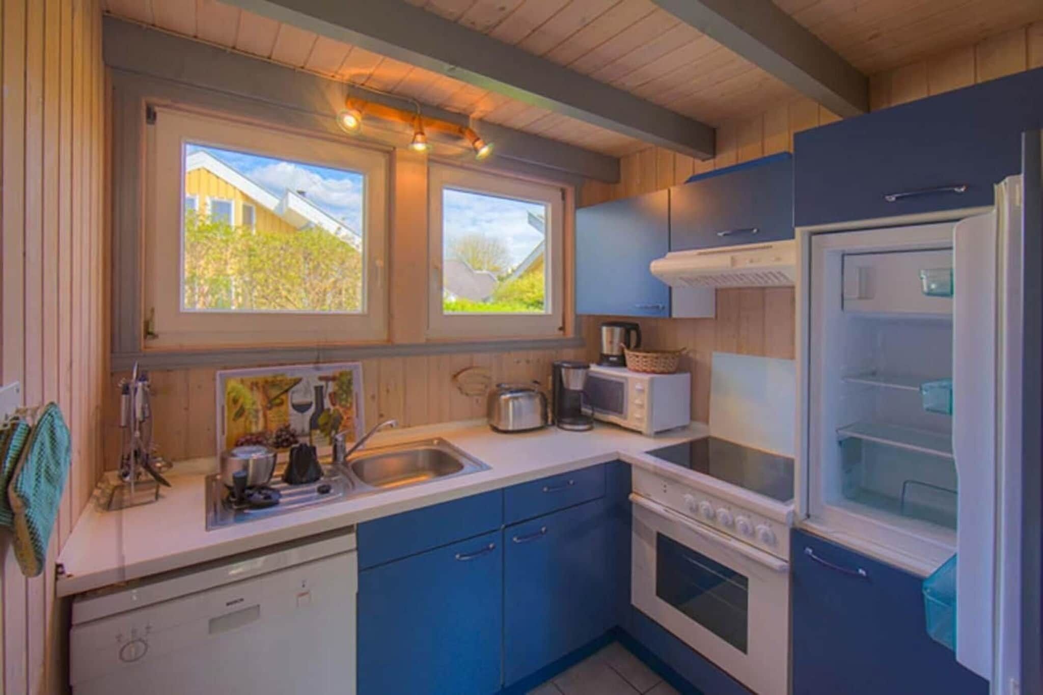 Una cocina compacta con gabinetes azules, un refrigerador blanco, un microondas y una estufa. Hay dos ventanas que ofrecen luz natural y vistas al exterior.