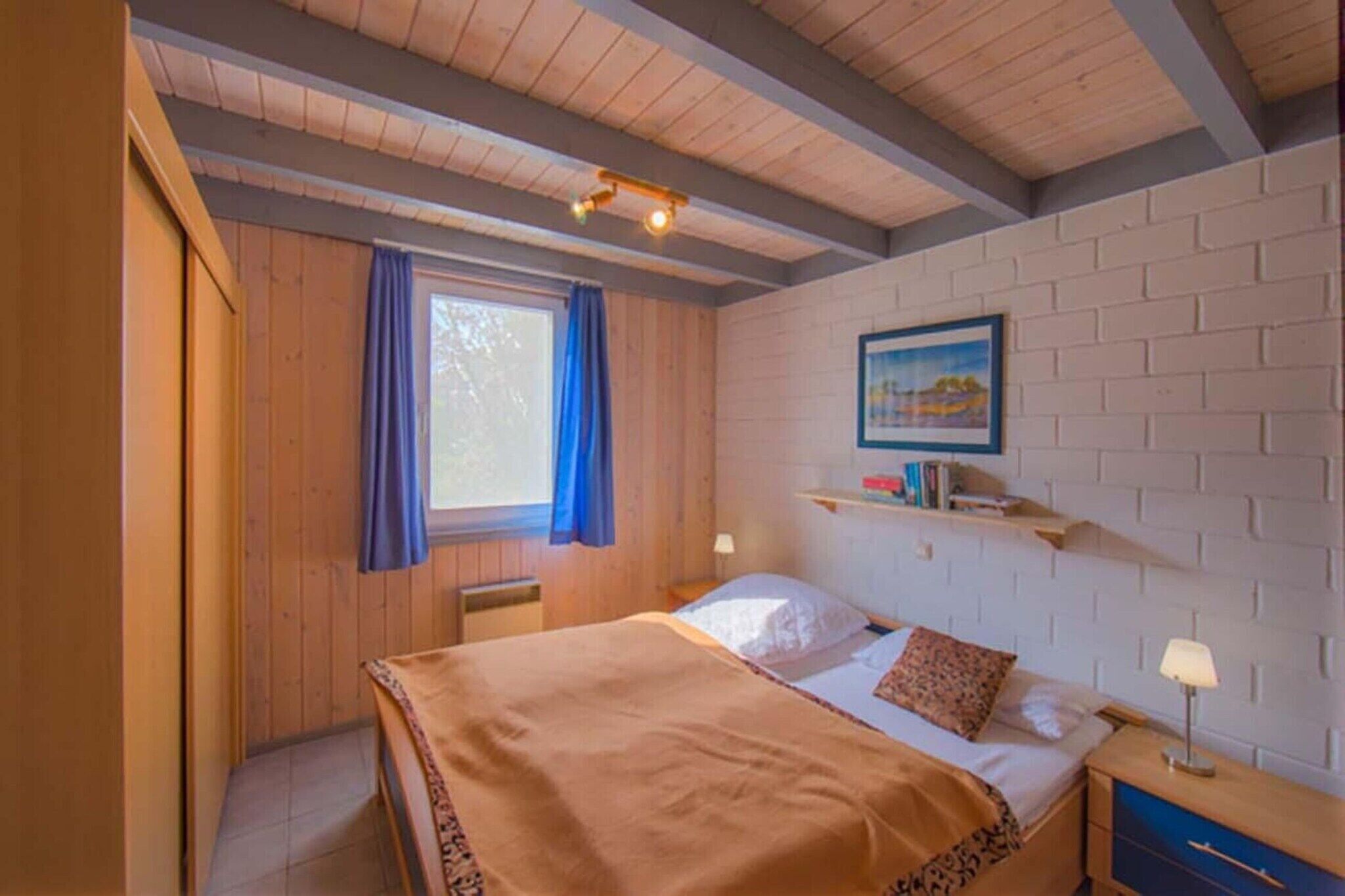 A cozy bedroom with a wooden ceiling, a bed with a brown blanket, a nightstand with a lamp, and a window with blue curtains.