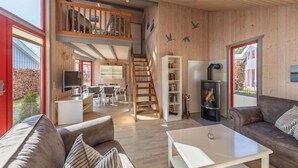 A spacious living area with a high ceiling, wooden walls, a staircase to an upper level, a fireplace, and a large window offering a view of the outdoors.