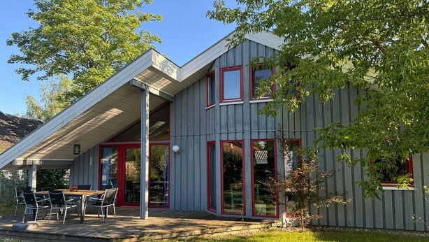 Ein modernes Haus mit überdachter Terrasse, roten Fensterrahmen und einem kleinen Garten.