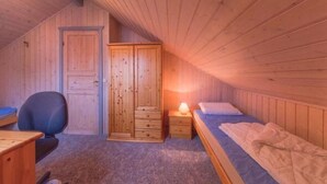 Une petite pièce chaleureuse aux murs en bois et au plafond en pente. Elle comprend un lit, un bureau, une chaise et une armoire.