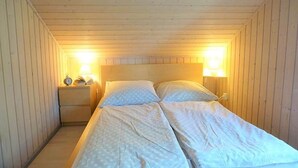 Una cama individual con cabecera de madera en una habitación estrecha con paredes y techo inclinados de madera. Hay dos lámparas de noche y una mesita de noche pequeña con un reloj.