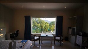 A hotel room with a bed, a desk, two chairs, a TV, and a window with a view of greenery. - Hotel Garnet (Bokakhat)