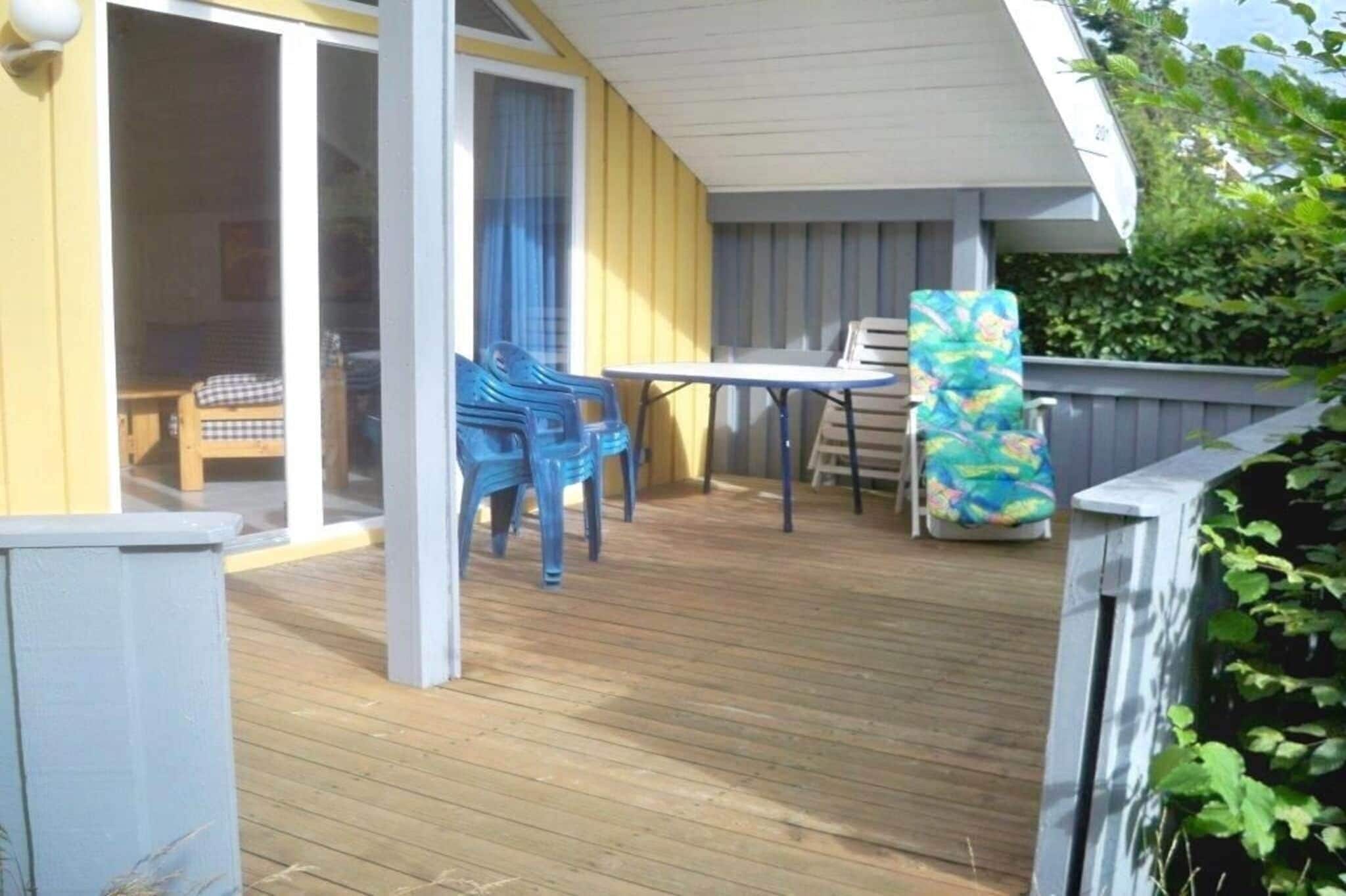 Una terraza de madera con una mesa y sillas, un ventanal corredero y un área cubierta con un banco.