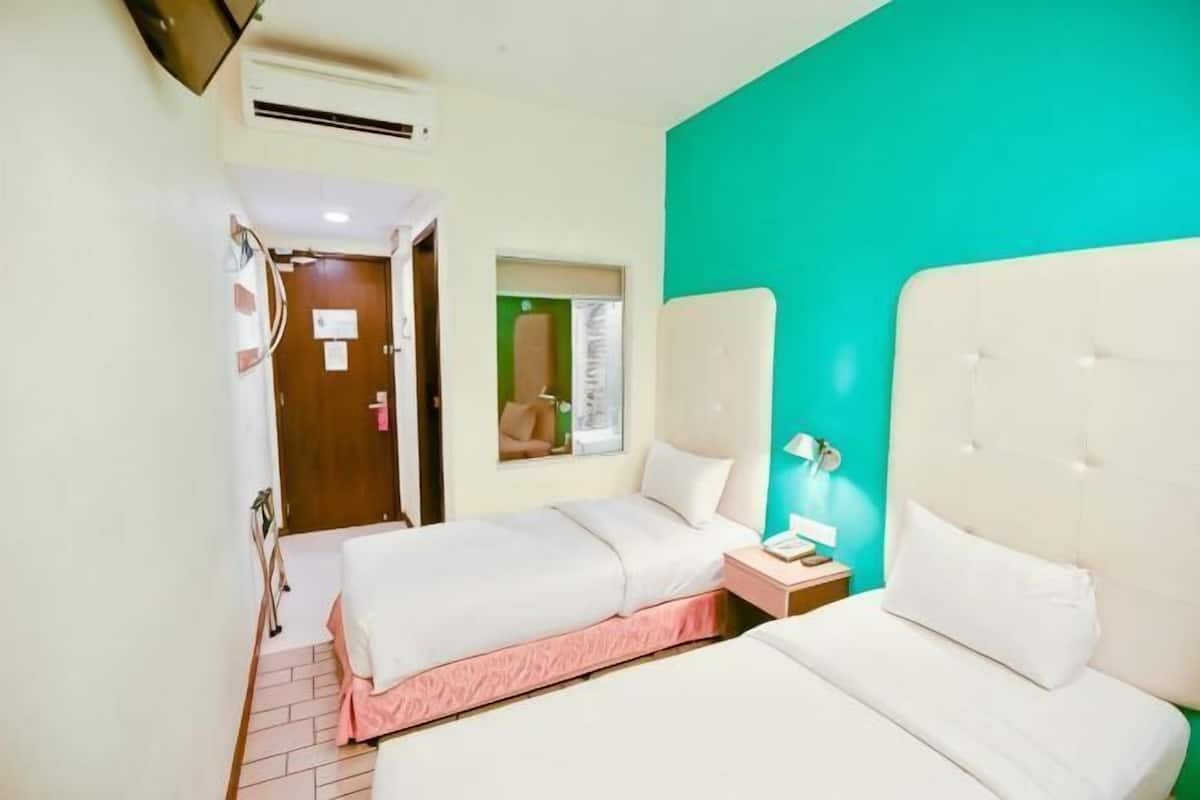 A hotel room with two beds, a window, a door, and a small bedside table with a lamp.