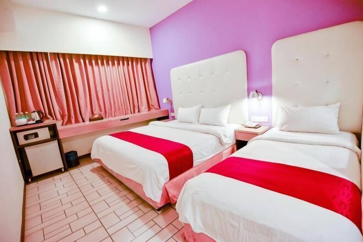 A hotel room with two beds, a nightstand, a window with curtains, and a tiled floor.