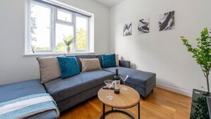 A modern living room with a grey sectional sofa, a small round table with a bottle and glasses, and a window with a view of greenery. - Ealing 3 Bath Flat w/ Parking | 10 mins to Station (London)