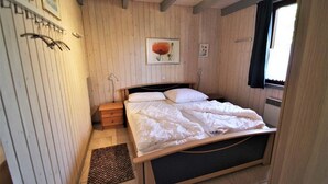 A bedroom with a wooden bed frame, a bedside table, a framed picture on the wall, and a window with curtains. - Ferienhaus im Ferienpark Mirow (Granzow)