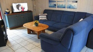 Un salon avec un canapé bleu, une table basse en bois sur laquelle est posé un vase, un téléviseur sur un meuble en bois, et un mur orné de deux tableaux.