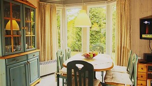 Un comedor con ventanal panorámico, una mesa con sillas, un mueble con puertas de cristal y un televisor sobre un soporte.