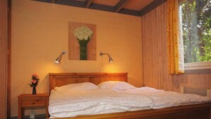 Una cama de madera con ropa de cama blanca, dos lámparas de noche, una mesita de noche con un florero y una ventana con cortinas.