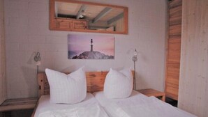 Un dormitorio con cabecera de madera, cama con ropa blanca, mesita de noche con lámpara, espejo en la pared e imagen de un faro.