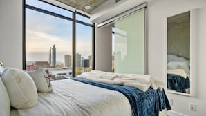 Un dormitorio con una cama grande, vistas a la ciudad a través de ventanales del suelo al techo y un espejo grande en la pared.