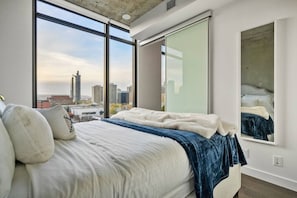 Quarto com uma cama grande, vista da cidade através de janelas do chão ao teto e um espelho grande na parede.