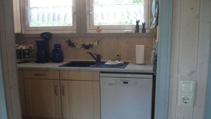 A compact kitchen with wooden cabinets, a white dishwasher, a sink, and a window with a view of greenery. - exklusives skandinavisches Ferienhaus (Granzow)