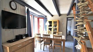 Un área de comedor con sillas de madera y una mesa, un mueble de madera con un televisor, y una estantería.