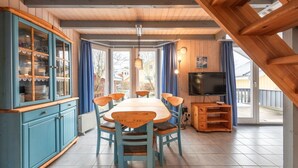 Un comedor con una mesa y sillas de madera, un armario azul con puertas de cristal, un televisor y vistas amplias a través de grandes ventanales.