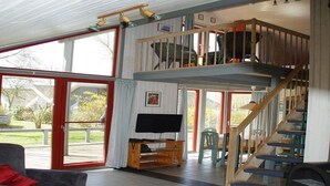 Amplia sala de estar con un gran ventanal corredero que da a una terraza exterior, una escalera de madera y un televisor sobre un mueble de madera.