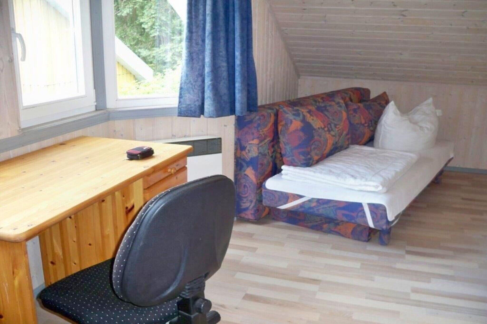 A room with a wooden desk, a black office chair, a bed with colorful bedding, and a window with curtains.