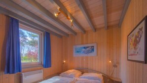 Une pièce de style cabane en bois, avec un lit, une fenêtre avec des rideaux, un tableau encadré au mur et une petite table avec une chaise.