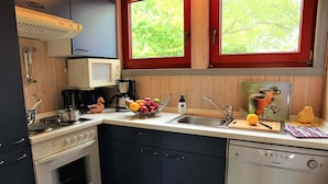 Una cucina compatta con piano cottura, forno, microonde ed elettrodomestici. Sopra il lavello c'è una finestra e una ciotola di frutta sul bancone.