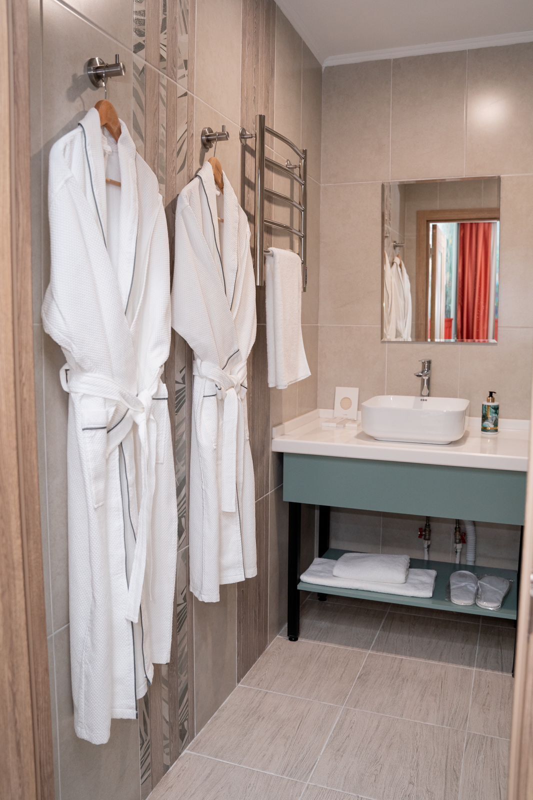 A modern bathroom with two white robes hanging on a rack, a sink with a mirror above it, and a towel rack.