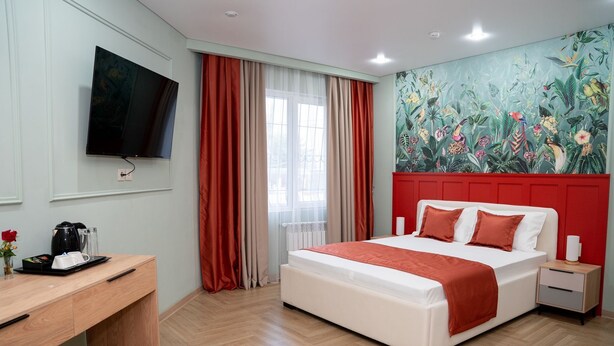 A hotel room with a bed, bedside table, a desk with a coffee maker, a TV mounted on the wall, and a mural with tropical plants. - Terrenkur boutique-hotel (Almaty)