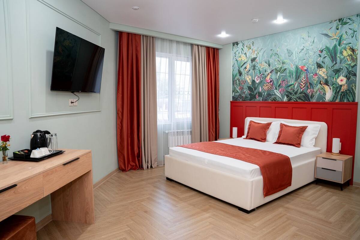 A hotel room with a bed, bedside table, a desk with a coffee maker, a TV mounted on the wall, and a mural with tropical plants.